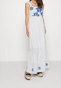Vrouw draagt een witte maxi-jurk met blauwe bladpatronen, mouwloos ontwerp, gelaagde zoom en sleehak sandalen tegen een effen achtergrond.