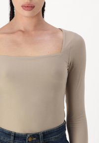 Helle beige Langarm-Bodysuit mit quadratischem Ausschnitt, aus glattem, dehnbarem Stoff, kombiniert mit dunklen Jeans mit hoher Taille.