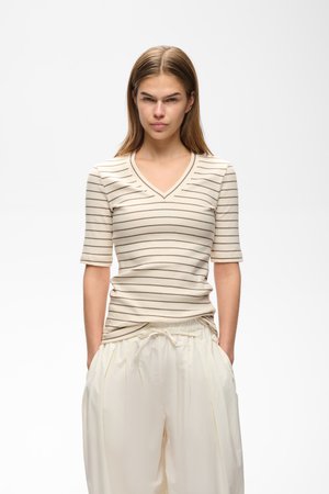 Tânără cu păr drept, castaniu deschis, purtând un tricou cu mânecă scurtă în dungi bej și maro, cu decolteu în V, și pantaloni largi crem, mâinile în buzunare.