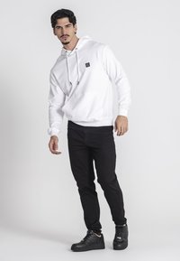 Biela mikina s kapucňou a predným vreckom, skombinovaná s čiernymi džínsami a čiernymi teniskami, predstavujúca ležérny outfit.