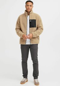 Tan fleece jacket with a black zippered chest pocket, high collar, and black zipper details. Paired with gray pants and brown sneakers.