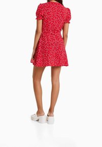 Červené mini šaty s bílým květinovým vzorem, s krátkými puff rukávy a zúženým pasem. Nosí se s bílými muly na blokovém podpatku. Hladká textura.