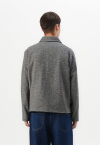 Grå fiskebensmønstret jakke med krage og lange ærmer, med et struktureret stof. Kombinert med blå denim bukser.