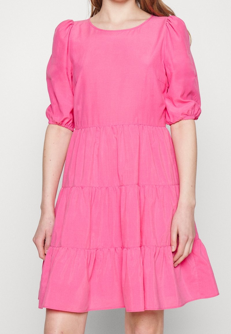 Femme portant une robe rose vif à étages avec des manches courtes bouffantes et un col rond, debout devant un fond clair uni.