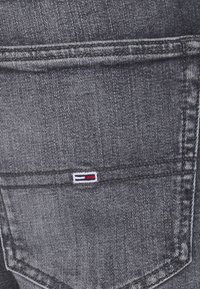 Šedé džíny z denimu s malým vyšitým logem v červené, bílé a modré barvě u zadní kapsy. Má klasické švy a design kapes.