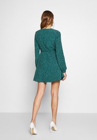 Vestido verde con lunares blancos, de mangas largas, con cintura entallada y falda acampanada. Llevado con tacones altos blancos, mostrando las piernas.
