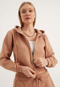 Felpa con zip color marrone chiaro, con cappuccio, tasche frontali, polsini a costine e cintura con cordino abbinata, realizzata in materiale morbido.