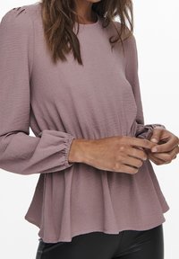 Haut à manches longues en tissu mauve doux à surface texturée, avec resserrement élastique aux poignets et taille froncée pour une coupe décontractée.