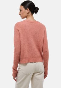 Pull rose, texturé, à manches longues et à la coupe légèrement courte. Poignets côtelés et col large; porté avec un pantalon clair.