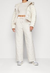 Kurtka puchowa i pikowane spodnie w jasnym kremowym kolorze. Kurtka posiada kaptur, wykończenia z prążkowanego materiału oraz teksturowaną tkaninę; spodnie mają elastyczną talię.