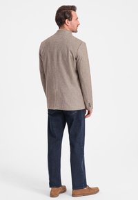 Brauner strukturierter Blazer, mit einer Schlitze hinten, kombiniert mit dunkelblauen Jeans. Modell trägt beigem Schuhe. Subtiler Schnittdetails an den Ärmeln.