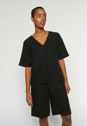 Frau mit kurzen Haaren, die ein schwarzes V-Ausschnitt-Kurzarmshirt und passende kniehohe Shorts trägt, vor einem einfarbigen Hintergrund stehend.