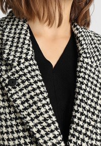 Abrigo de tweed con patrón de pata de gallo en blanco y negro, con cuello muesca y cierre de un solo botón, sobre una blusa negra de cuello en v.