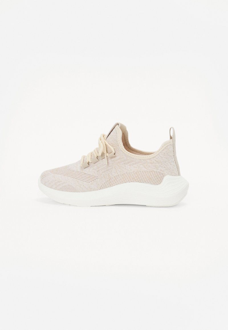 Chaussure de sport beige avec une tige en mesh texturé, des lacets et une épaisse semelle en caoutchouc blanc ; elle possède une languette au talon pour un enfilage facile.