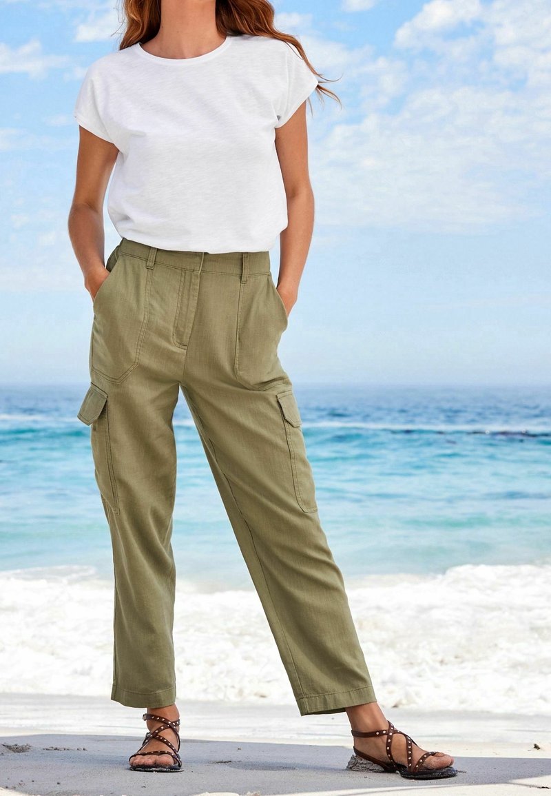 Femme debout sur la plage portant un t-shirt blanc, un pantalon cargo vert olive avec des poches, et des sandales brunes à fines lanières, avec l'océan en arrière-plan.