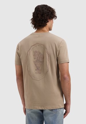 Man met krullend haar die een beige T-shirt draagt met een geborduurd design van een bloemenvaas en de tekst "Pure Paths" op de achterkant, gecombineerd met lichtblauwe spijkerbroek.