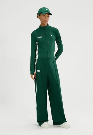 Groene sportset met een ritssluiting jas met witte zijkanten en bijpassende brede broek, aangevuld met een groene pet en witte sneakers.