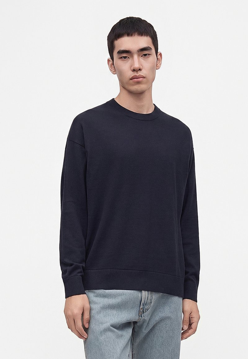 Jeune homme aux cheveux courts portant un pull noir uni à manches longues et un jean bleu clair, debout devant un fond blanc.