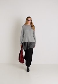 Un pull en tricot gris superposé sur un top plissé noir, associé à un pantalon noir ample et un sac rouge brillant, complété par des bottines noires.