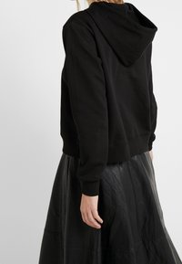 Felpa nera corta con cappuccio, caratterizzata da maniche lunghe e polsini a costine, abbinata a una gonna di pelle nera con cuciture in vista.