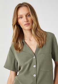 Camisa de manga corta en verde olivo, hecha de una tela suave, con cuello en V, cinco botones y un bolsillo en el pecho. Textura sutil en el material.