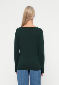 Pull en mailles texturées vert foncé avec un large décolleté et des manches longues, associé à un jean en denim bleu clair et ample.
