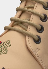 Κοντινό πλάνο ενός μπεζ δερμάτινου παπουτσιού με μπεζ κορδόνια περασμένα από μαύρες μεταλλικές τρύπες και πράσινη κεντημένη φυτική διακόσμηση στα πλάγια.