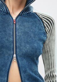 Närbild på en person som bär en blå stickad zip-up-jacka med en röd dragkedjekäpp och ribbade ärmar med vita och svarta ränder.