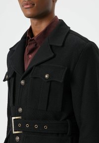 Zwarte, geribbelde jas met twee bovenzakken, metalen knopen, een kraag en een tailleband met ceintuur, gecombineerd met een bordeauxrode overhemd eronder.