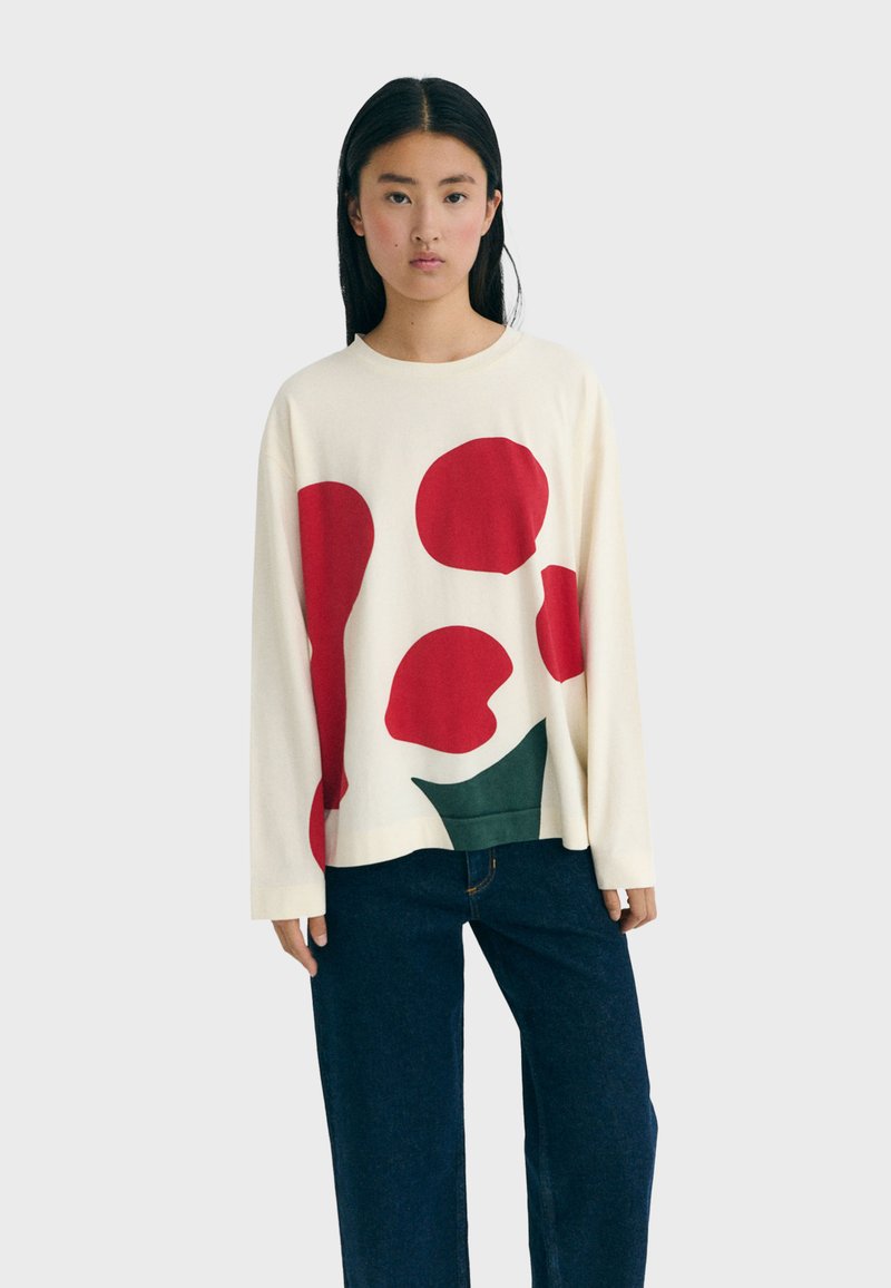 Woman with long black hair wearing a white sweater with large red and green abstract shapes and dark blue wide-leg jeans.