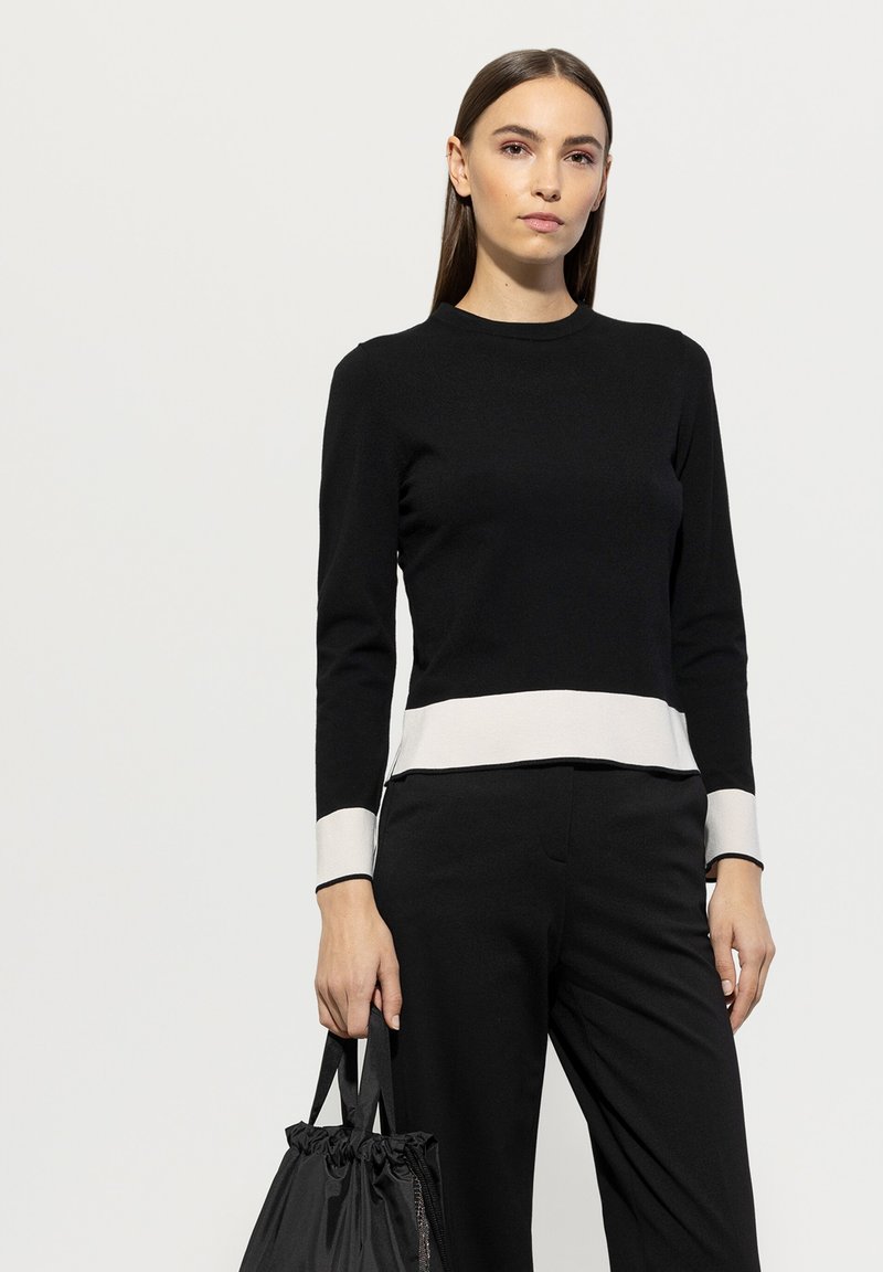 Schwarzer Pullover mit langen Ärmeln, ausgestattet mit einem kontrastierenden weißen Saum und Bündchen. Getragen mit einer schwarzen Hose und einer Zugbandtasche.