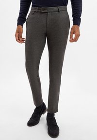Graue, strukturierte maßgeschneiderte Hosen mit schlankem Schnitt, ausgestattet mit einem Knopfverschluss, Gürtelschlaufen und Seitentaschen. Getragen mit dunklen Schuhen.