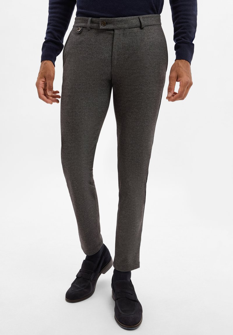 Graue, strukturierte maßgeschneiderte Hosen mit schlankem Schnitt, ausgestattet mit einem Knopfverschluss, Gürtelschlaufen und Seitentaschen. Getragen mit dunklen Schuhen.