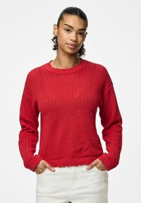Maglione rosso lavorato a maglia con un motivo testurizzato, scollatura a onde e polsini a coste. Il modello indossa pantaloni bianchi con le mani nelle tasche.