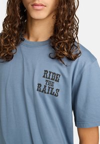 Person med langt krøllet hår iført en blå T-shirt med sort tekst, der står "RIDE THE RAILS" på brystet.