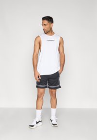 Λευκό αμάνικο αθλητικό τοπ με το λογότυπο "UNDER ARMOUR", σε συνδυασμό με μαύρο σορτς με λευκές λεπτομέρειες. Φορεμένο με λευκές κάλτσες και μαύρα αθλητικά παπούτσια.