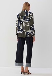 Lange mouwen, gebloemde blouse in donkergroen, marineblauw en crème. Gecombineerd met wijde donkerdenim broek met grijze boorden en zwarte haksandalen.