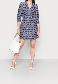 Niebieska sukienka w wzory z dekoltem w kształcie litery V i rękawami 3/4 z bufkami. W komplecie z białymi sandałami na obcasie i białą torebką.
