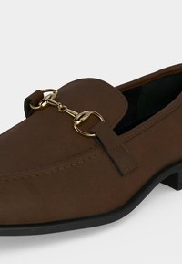 Mocassin en cuir marron avec semelle noire et détail en métal doré en forme de mors de cheval sur le dessus, photographié sur fond blanc uni.