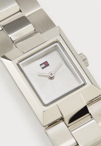 Silver rectangular watch with a brushed metal strap and a polished case. The face has a silver background with a small logo and thin hands.