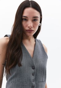 Gilet a spina di pesce grigio con profondo scollo a V, chiusura con bottone e due tasche frontali. Tessuto liscio e leggermente lucido. Il modello ha i capelli lunghi e indossa orecchini.