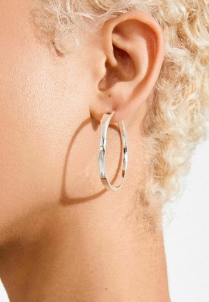 Silberner Creolen-Ohrring mit gedrehtem Design, reflektierender Oberfläche und mittlerer Dicke, getragen zu einer kurz geschnittenen Frisur auf der Haut.