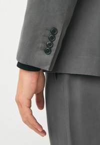 Grauer Anzugjacke aus texturiertem Stoff, mit vier schwarzen Knöpfen an den Bündchen und einem sichtbaren schwarzen Strickdetail am Ärmelabschluss.