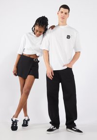 White oversized t-shirt with a small black logo, paired with a black wrap skirt featuring a silver buckle. Black sneakers complete the look.