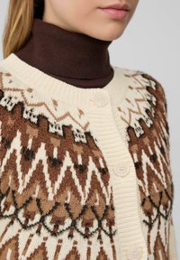 Sweter kardigan w kolorze kremowym z brązowymi geometrycznymi wzorami; posiada prążkowany kołnierz, trzy guziki oraz miękkie, teksturowane wykończenie.