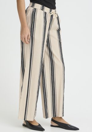 Frau trägt beige, weit geschnittene Hosen mit schwarzen vertikalen Streifen und ein schwarzes Oberteil, kombiniert mit schwarzen spitzen Slingback-Schuhen.