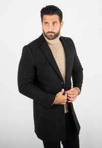 Manteau en mélange de laine noir à coupe ajustée, fermeture à un bouton, et revers crantés, superposé sur un pull en tricot torsadé beige.