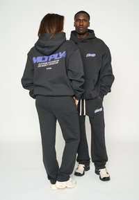 Felpe e pantaloni sportivi neri realizzati in materiale morbido, con loghi stampati viola. Il design è a vestibilità comoda e presenta tasche frontali e cordini.