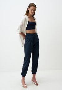 Navyblaue, hoch taillierte Hose mit Falten, kombiniert mit einem enganliegenden dunklen Oberteil und einem lockeren weißen Hemd mit aufgerollten Ärmeln und offener Vorderseite.