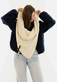 Gruba, kremowa dziana chusta w kształcie trójkąta z teksturowanym wzorem, akcentowana brązową skórzaną metką i regulowanym sznurkiem.